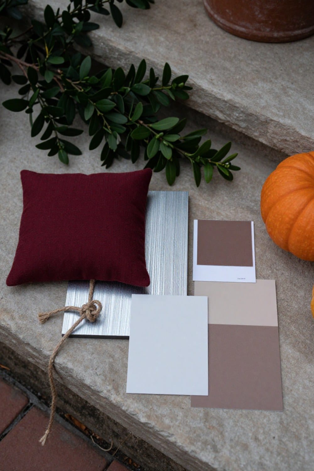 A flat-lay of an autumn patio color palette -- oxblood fabric, silvered teak, jute-look rope, and deep green boxwood -- with no orange in it
