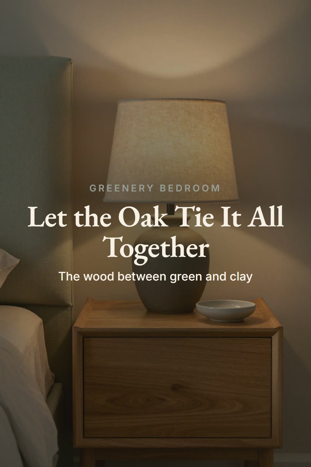 A honey oak nightstand with a clay lamp and oatmeal linen shade beside a sage headboard in lamplight