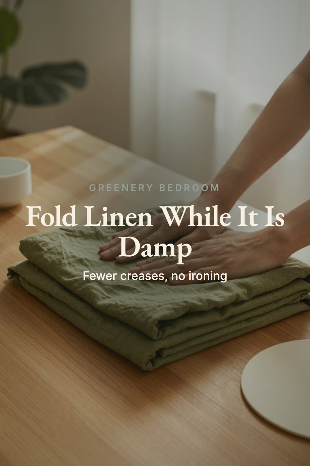 Hands folding a slightly damp olive linen sheet in thirds on a wooden table and pressing it flat
